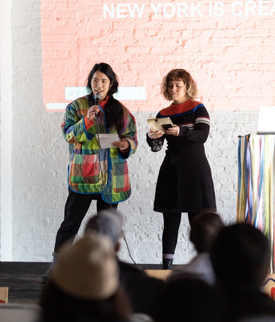 Photo of Annie speaking into a microphone in a multi-colored jacket at a New York is Creative event. Alexa stands next to her with an open book, while an audience looks on.