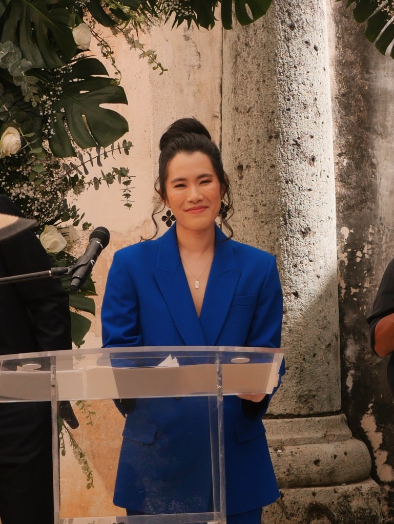 Photo of Annie in a blue suite at a clear podium officiating a wedding. Photo by Jenna Ruddock.