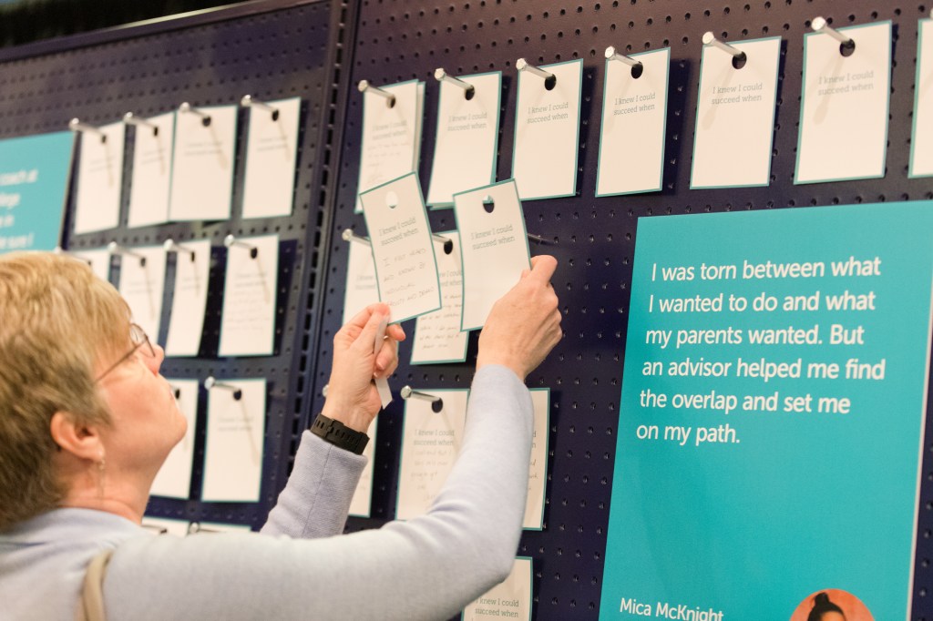 Photo of a participant placing a handwritten story of a turning point ontp a peg. There is an image to the right with a quote that says "I was torn between what I wanted to do and what my parents wanted. But an advisor helped me find the overlap and set me on my path."